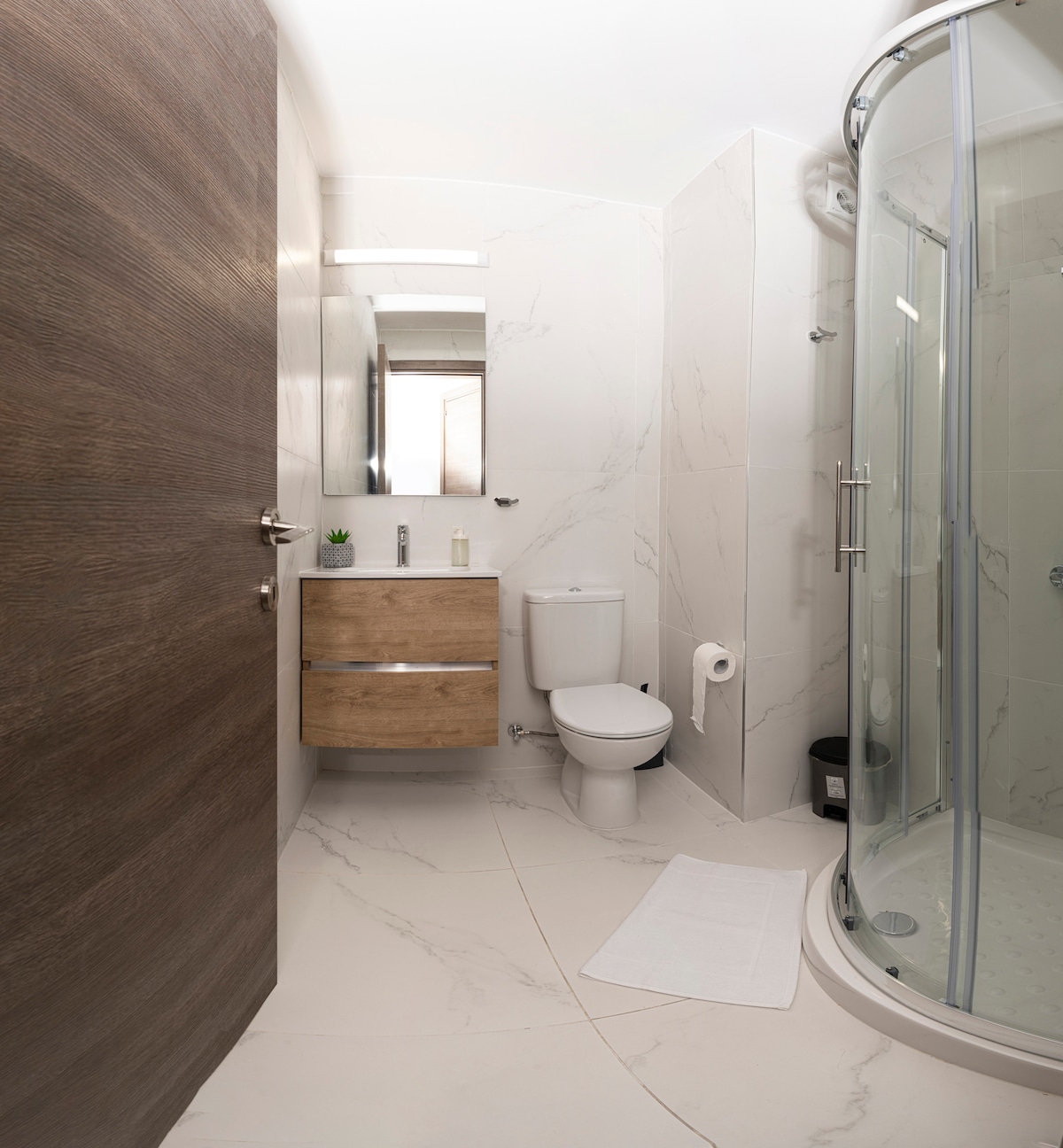 A modern bathroom features light marble tile and a glass shower enclosure. A wooden vanity with a curved design is paired with a mirror above. A white toilet and a small plant add subtle decor. Soft white towels are neatly arranged on the floor.