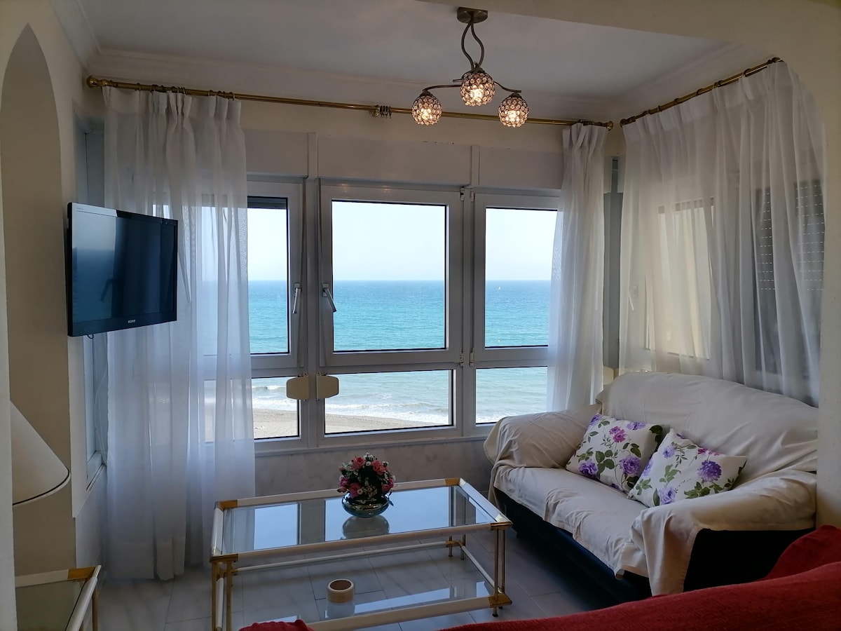 A cozy living area features a comfortable sofa adorned with floral accents. Large windows offer panoramic views of the sea, while sheer white curtains gently frame the view. A glass coffee table sits in front, and a television is mounted on the wall.