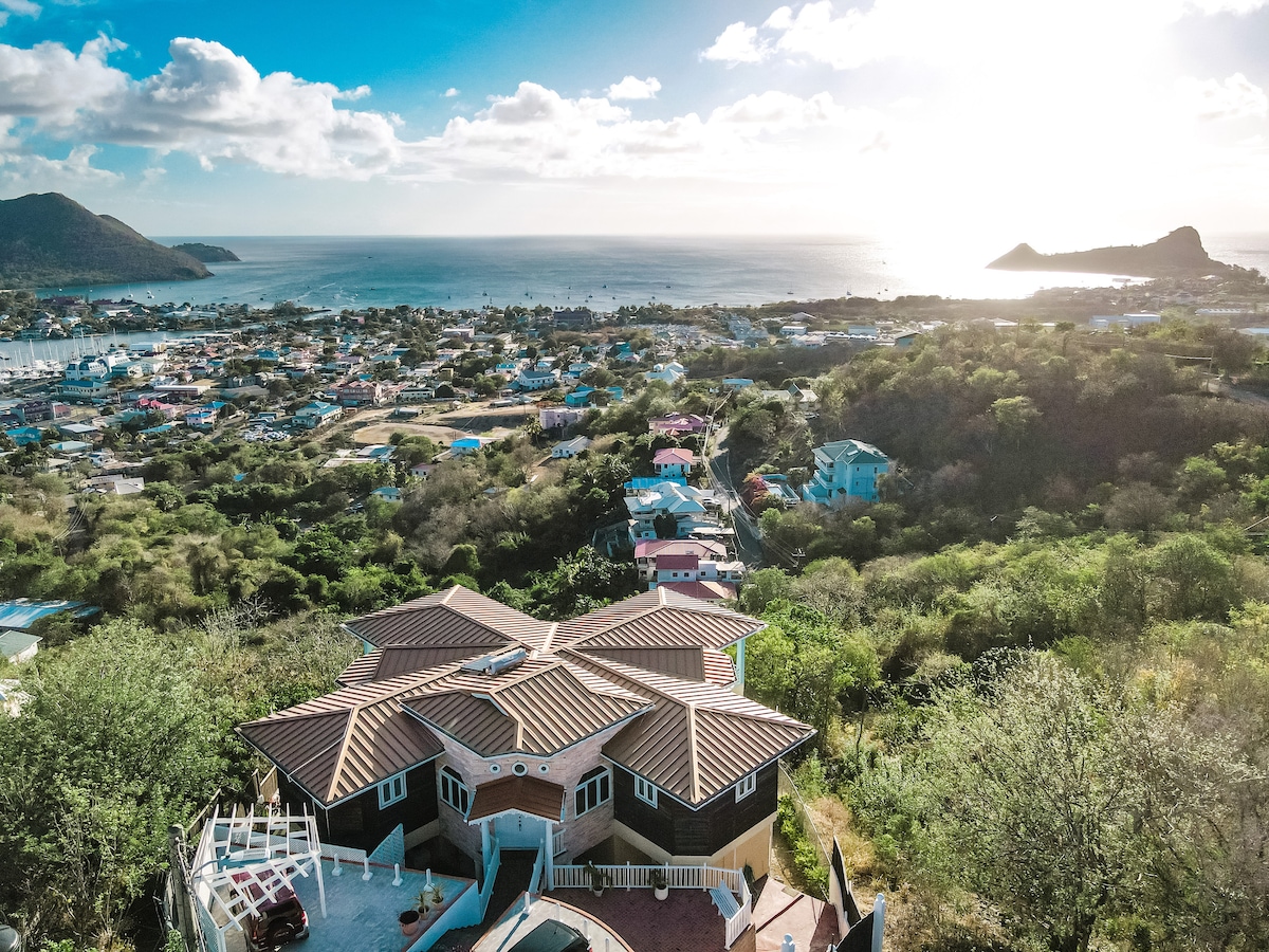 An aerial view of a spacious villa set amidst lush greenery, showcasing a unique octagonal shape. The surrounding landscape features colorful residential structures and panoramic views of Gros Islet, Rodney Bay Marina, and Pigeon Island under a bright sky.