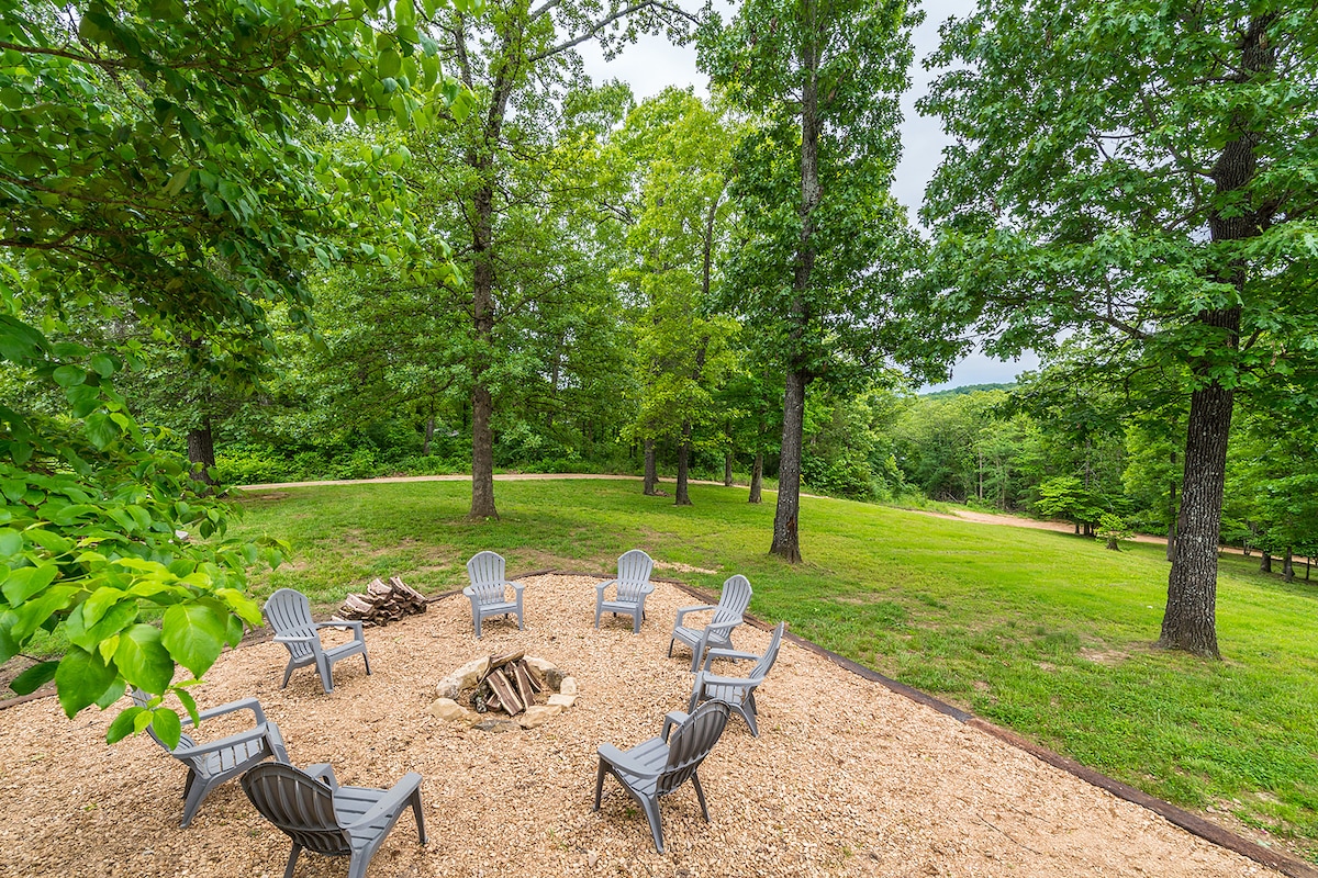 A fire pit area is surrounded by multiple gray lounge chairs positioned on a gravel surface. Lush greenery and tall trees create a natural backdrop, while an open grassy space offers room for relaxation and outdoor activities.