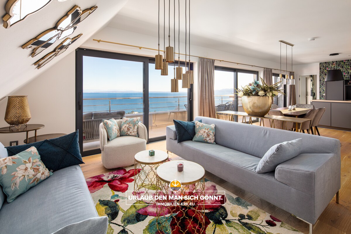 A modern living area is highlighted by large windows offering views of the Adriatic coastline. Elegant seating arrangements include a spacious grey sofa and two armchairs, complemented by decorative side tables. Stylish light fixtures hang from the ceiling, enhancing the contemporary decor.