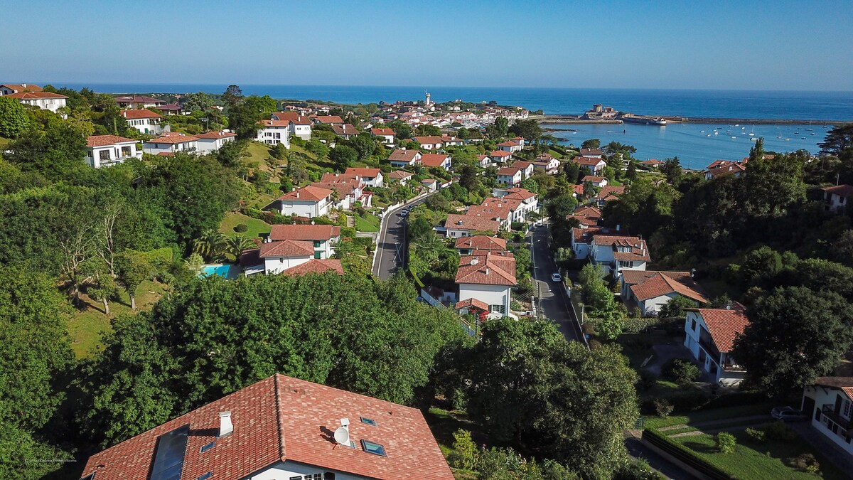 Aerial view of a coastal neighborhood featuring homes with red-tiled roofs and lush greenery. The ocean is visible in the background, alongside a distant view of the town and boats in the water. The scene captures a serene, picturesque landscape.