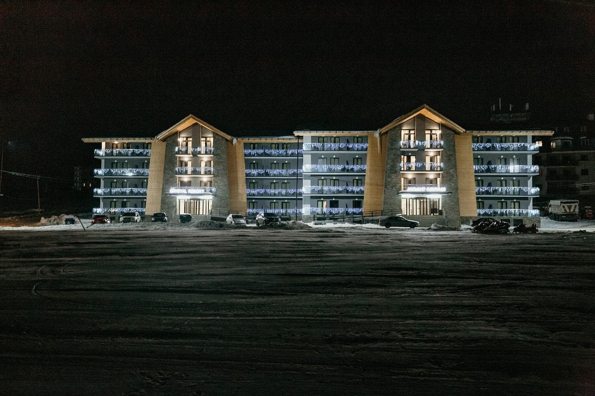 A three-story building with a distinctive wooden facade is illuminated at night, showcasing its multiple balcony areas. Snow-covered ground surrounds the structure, and several parked vehicles are visible in the foreground, indicating a winter setting near recreational facilities.