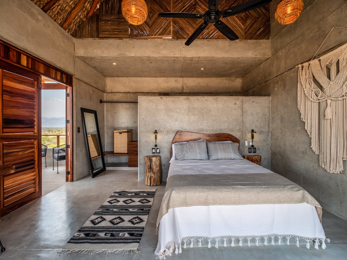 A modern bedroom features a spacious bed with neutral bedding, complemented by a decorative wall hanging. Natural light fills the room through an open door, where a wooden chair and table are visible. Unique lighting fixtures enhance the room's design.