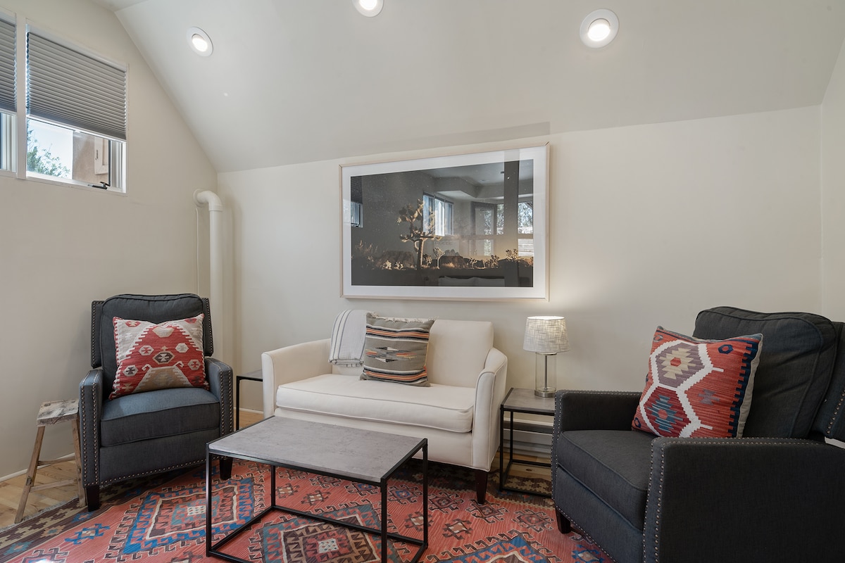 A cozy living area features two armchairs and a white sofa set against light-colored walls. A coffee table sits centered on a patterned rug. Natural light enters through a nearby window, and a piece of wall art adds a touch of character to the space.