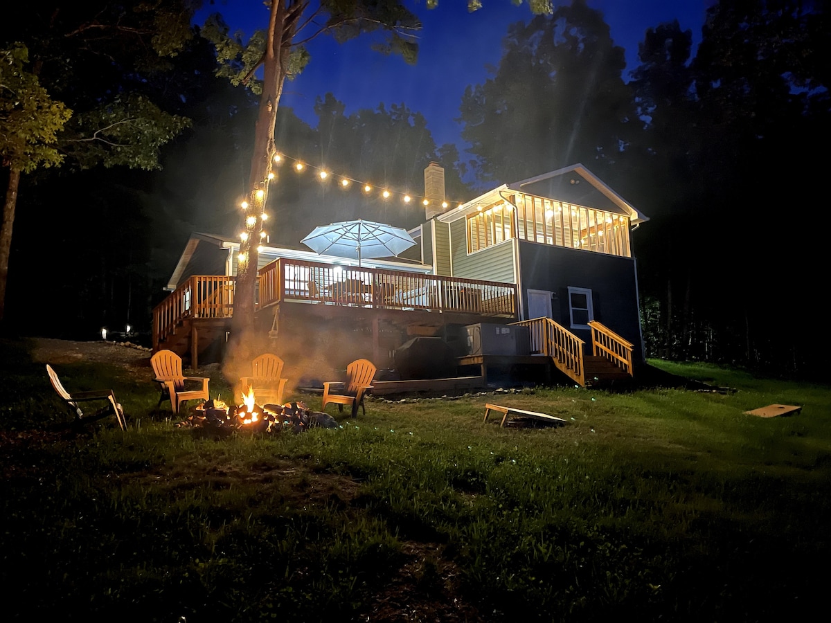 The property is illuminated by warm string lights, highlighting the expansive deck and cozy seating area. A fire pit sits in the foreground, surrounded by chairs. The home’s large windows glow softly, showcasing the inviting atmosphere of the retreat nestled in the trees.