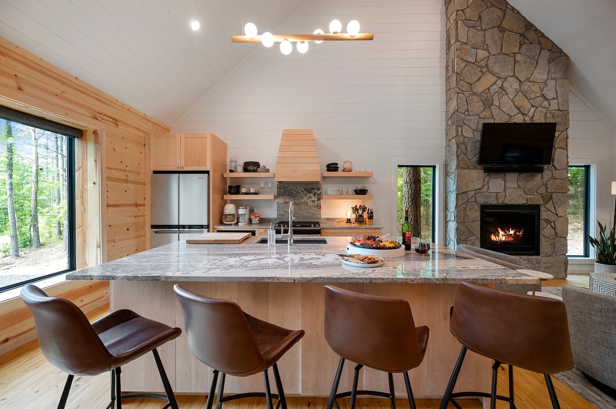 The modern kitchen features an oversized island with a sleek stone countertop, surrounded by four padded chairs. High-end appliances and minimalist cabinetry complement the stone fireplace, enhancing the cabin's contemporary design. Large windows provide views of the surrounding trees, inviting natural light into the space.