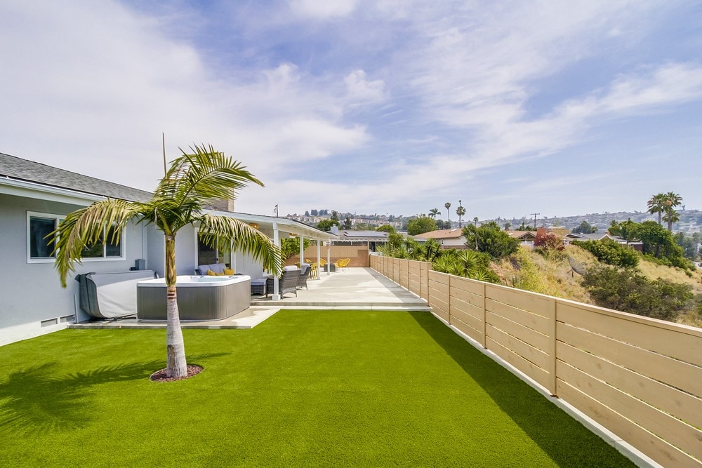 A spacious backyard is showcased, featuring a well-manicured lawn with artificial turf and a hot tub. Outdoor seating is arranged beneath a covered patio, providing a view of the surrounding landscape and distant mountains under a clear sky.