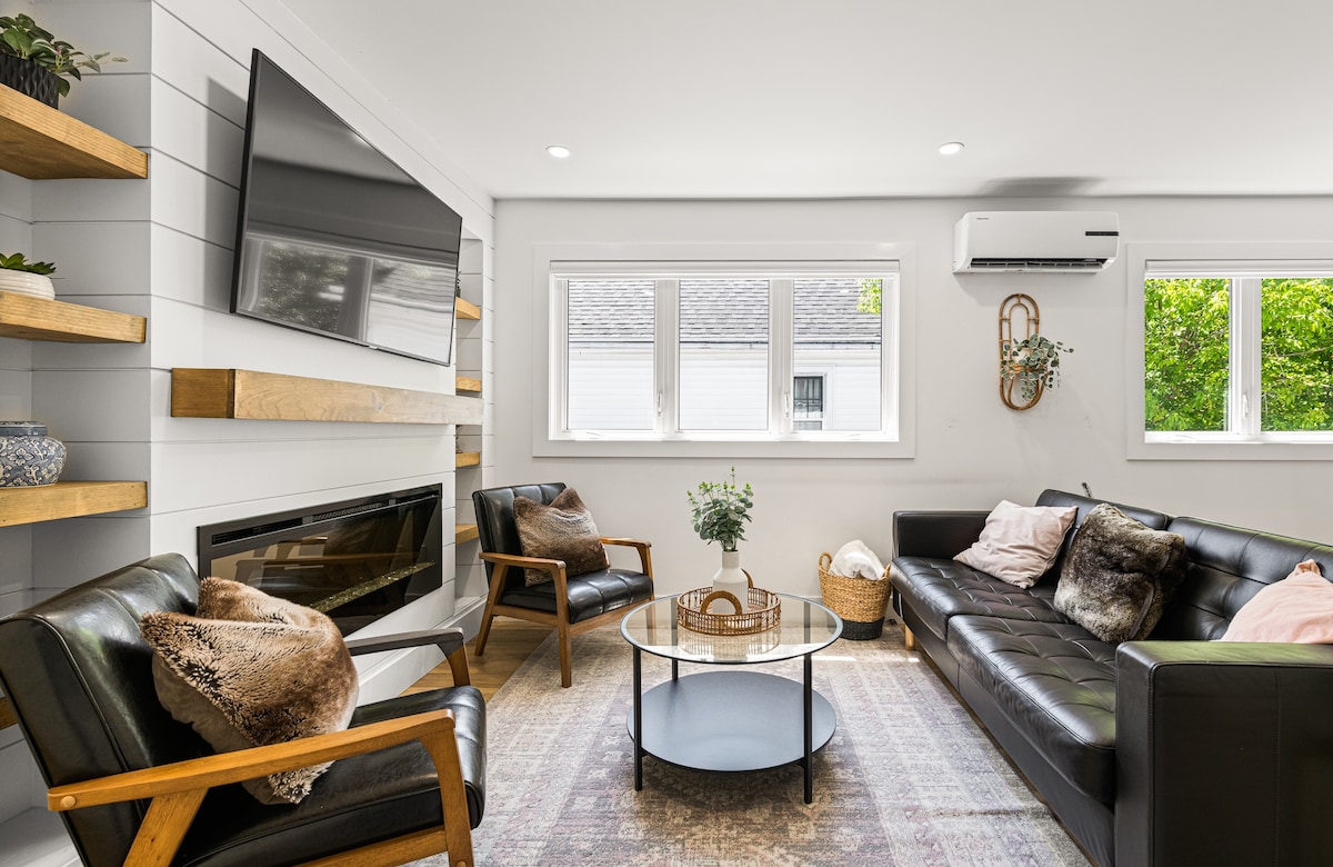 A cozy living area features a modern black sofa and two armchairs arranged around a circular coffee table. A wall-mounted television and an elegant fireplace are present, while natural light streams through the multiple windows adorned with minimalistic decor.