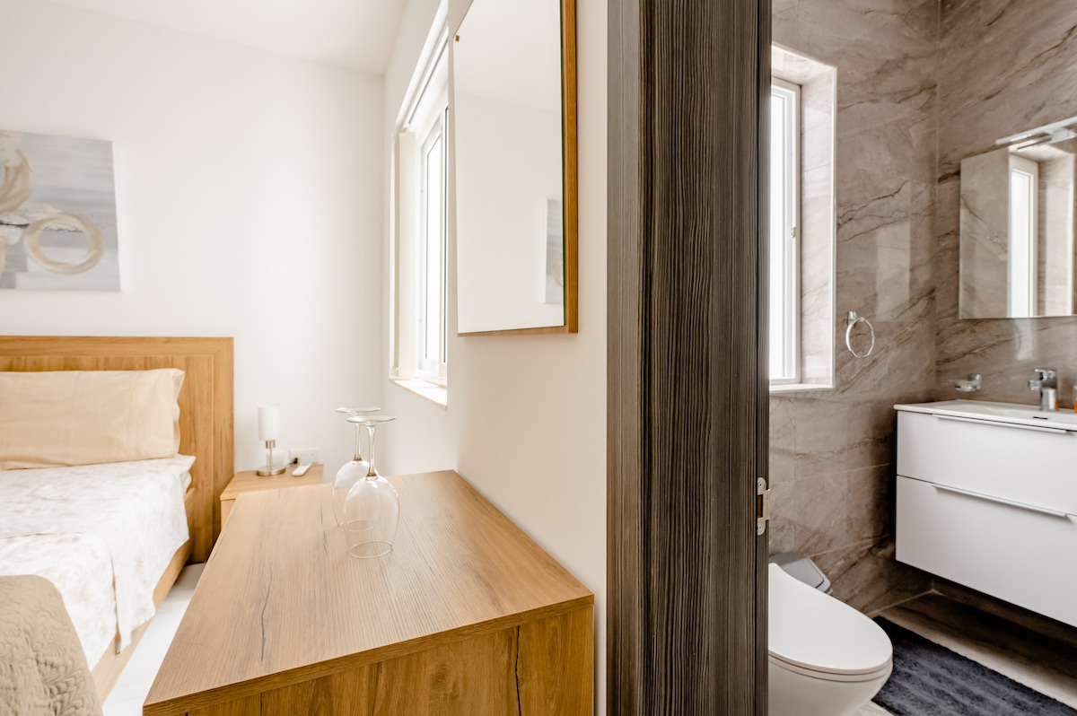 A double bedroom is featured, with a cozy bed positioned near a wooden dresser. Natural light enters through large windows. The adjoining private bathroom is visible, showcasing modern fixtures and a sleek design, with a neutral color scheme enhancing the area.