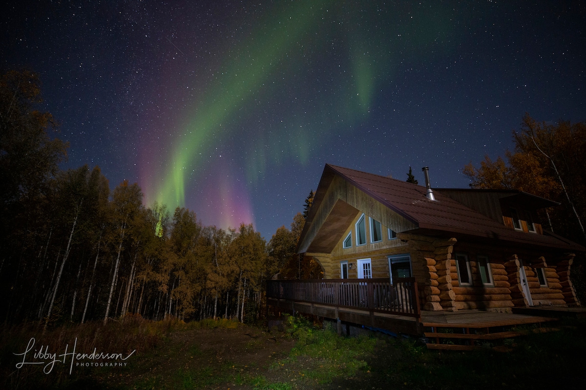 A custom log home is shown beneath a night sky illuminated by vibrant auroras. The structure features a wrap-around deck, and dense trees surround the property, enhancing the sense of tranquility in the natural setting.
