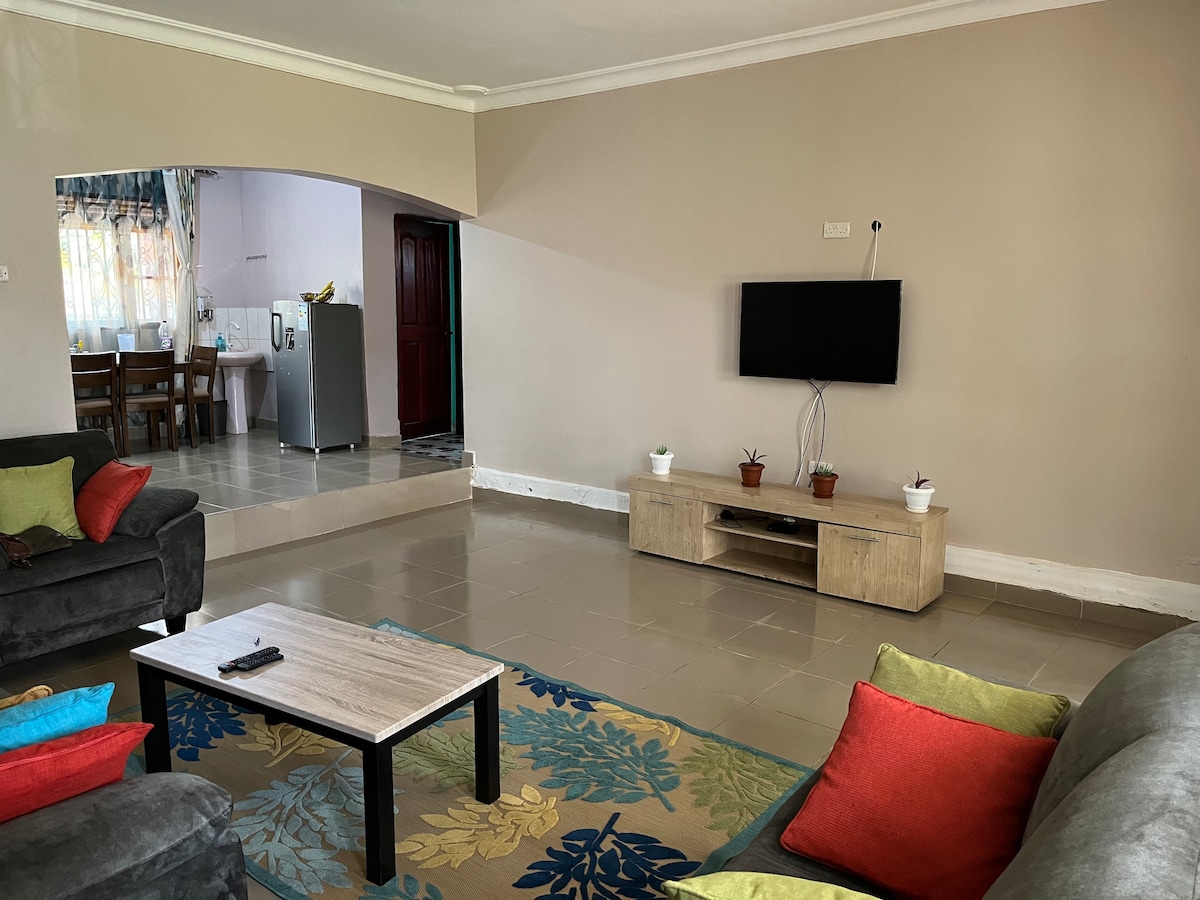 A spacious living area features a neutral wall color and a large carpet with floral patterns. Comfortable gray sofas are arranged around a small coffee table. A television is mounted on the wall, with potted plants adorning the entertainment unit. The dining area is visible in the background.