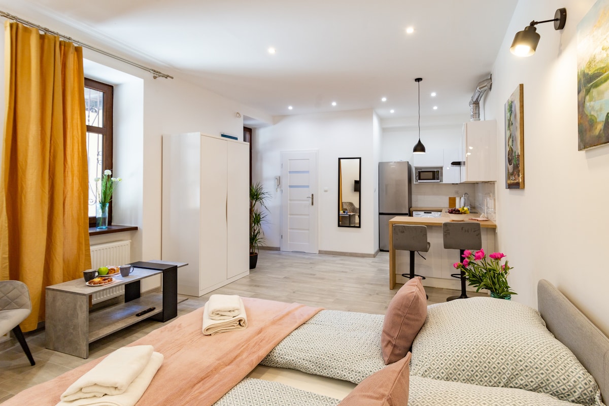 The studio apartment includes a spacious sleeping area with two beds, fresh linens, and decorative pillows. A modern kitchen is visible with essential appliances and seated dining at a countertop. Soft lighting enhances the warm hues of the curtains, while plants add a touch of nature.