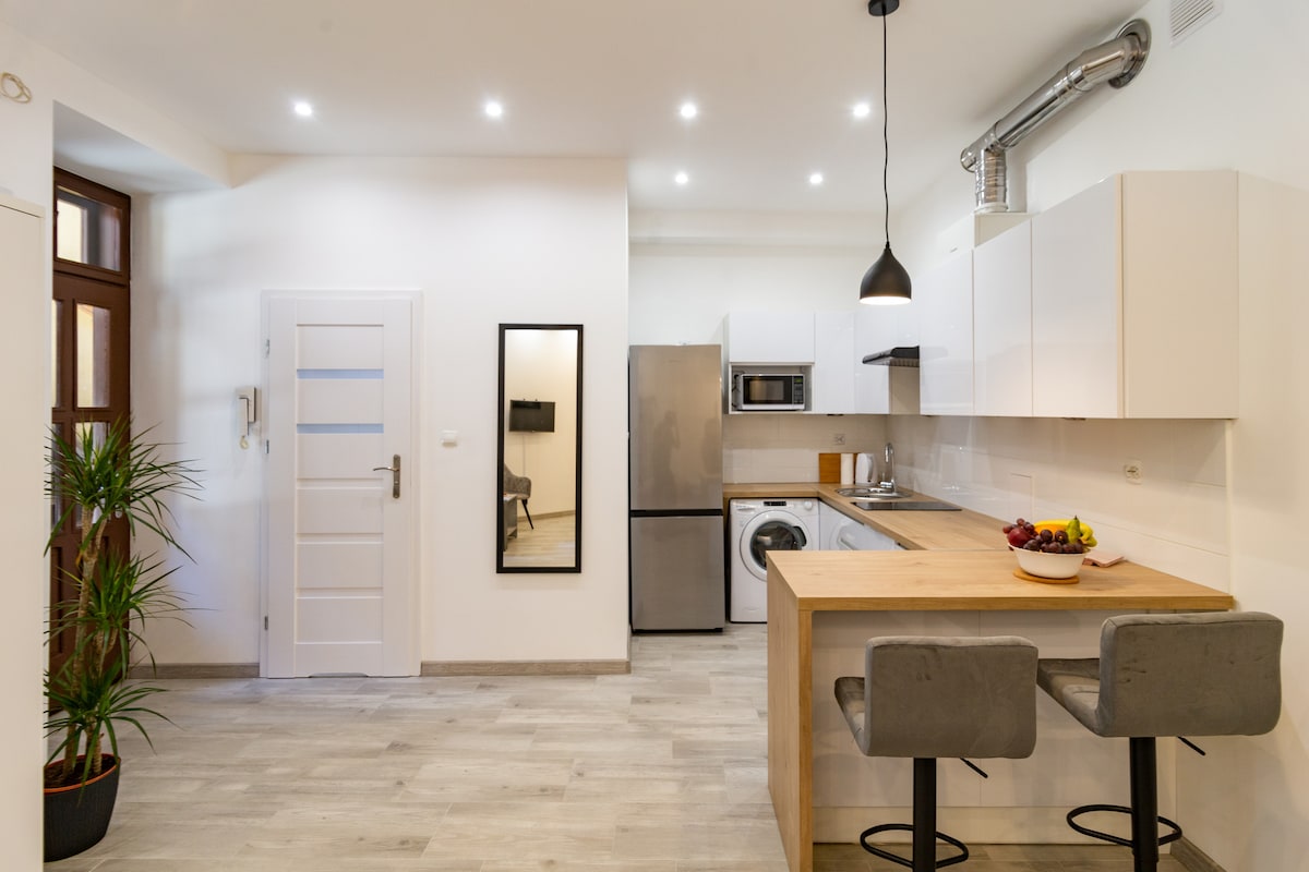 A modern kitchen area in the studio features sleek white cabinetry, stainless steel appliances, and a wooden dining table with two stools. A small plant adds a touch of greenery, while ample lighting brightens the space, creating a functional cooking environment.