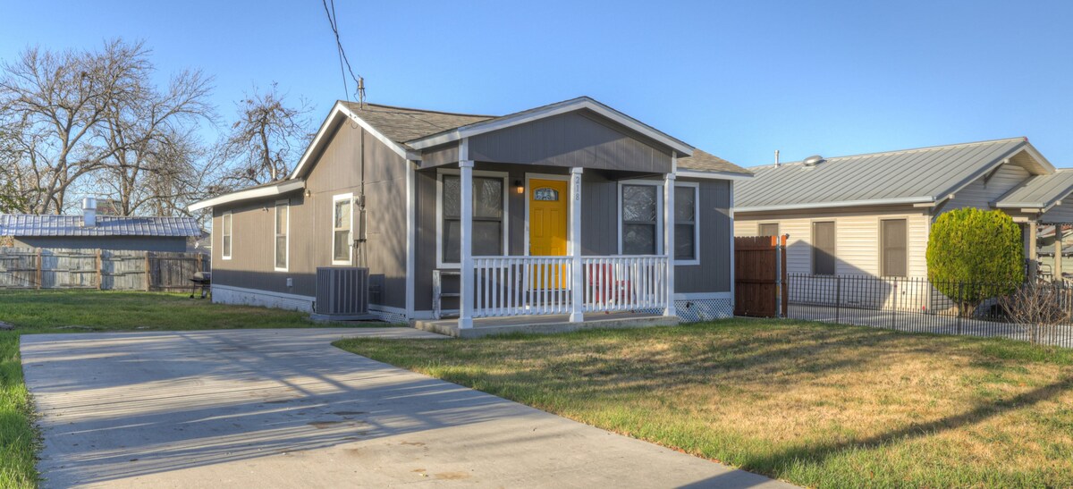 The exterior of a single-story home is shown, featuring a light gray facade with a welcoming yellow front door. A covered porch is visible, complemented by white railings. A lawn and driveway provide access, with neighboring homes visible on either side.