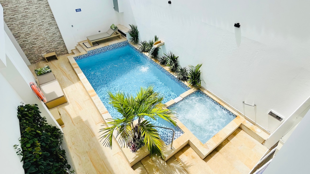 An outdoor pool area is surrounded by a sleek stone deck, featuring a refreshing swimming pool and a heated jacuzzi. Lush greenery, including palm plants, adds a tropical touch, while the sunlit space is complemented by a minimalist design and light-colored walls.