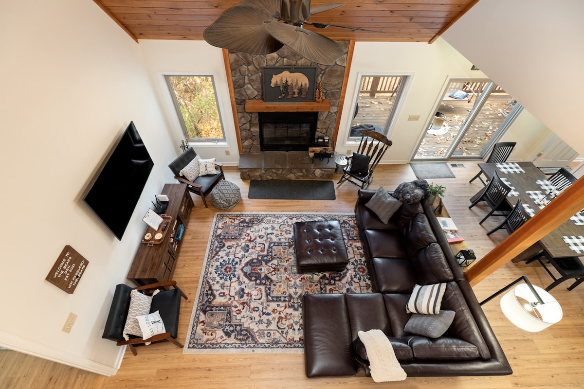 An aerial view of a spacious living area reveals a brown leather sectional couch positioned around a patterned area rug. A stone fireplace with a wooden mantle serves as a focal point, and large windows provide natural light. A dining table is visible in the background.