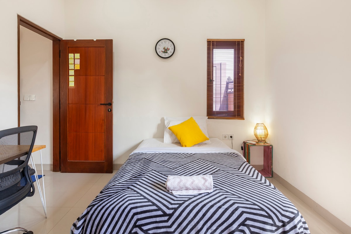 A cozy bedroom is presented, featuring a single bed with a striped blanket and a bright yellow pillow. A bedside lamp and a small side table add functionality. A window with wooden blinds allows natural light, while a clock is mounted on the wall above the bed.