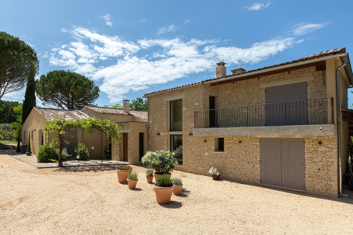 A stone house with a modern design features large windows and a balcony overlooks a peaceful landscape. The surrounding area is adorned with potted plants and gravel pathways, complemented by tall trees in the background under a partly cloudy blue sky.
