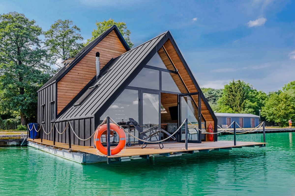 A modern floating house is presented on calm waters, featuring a unique triangular roof. Large glass windows allow natural light to fill the interior, while an orange life buoy adds a pop of color on the deck, surrounded by lush greenery.
