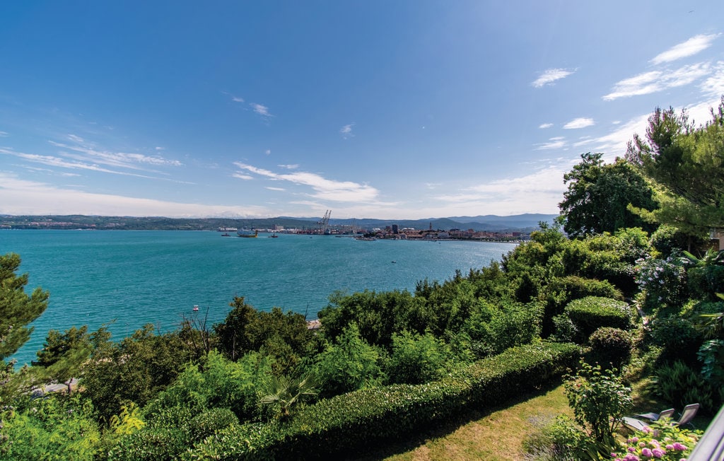 A serene coastal view showcases a vibrant blue sea meeting the horizon. Lush greenery and well-maintained hedges frame the landscape, while distant city structures and boats are visible in the water. Clear skies contribute to the tranquil ambiance.