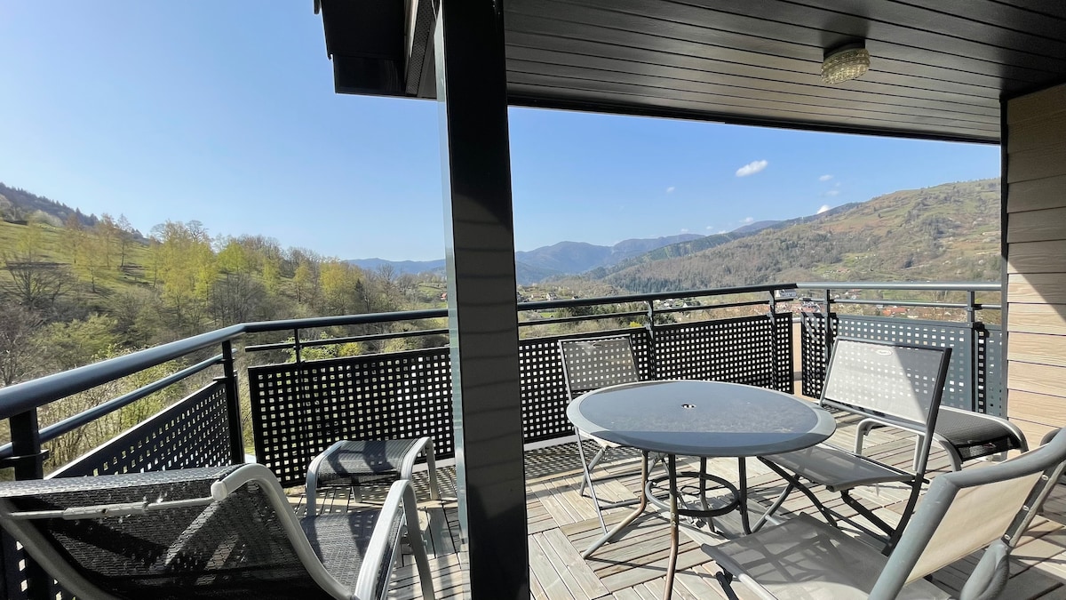 The balcony features a round table surrounded by six chairs, with two sun loungers positioned nearby. A scenic view of the valley and rolling hills is visible in the background, complemented by a clear blue sky and distant mountain ranges.