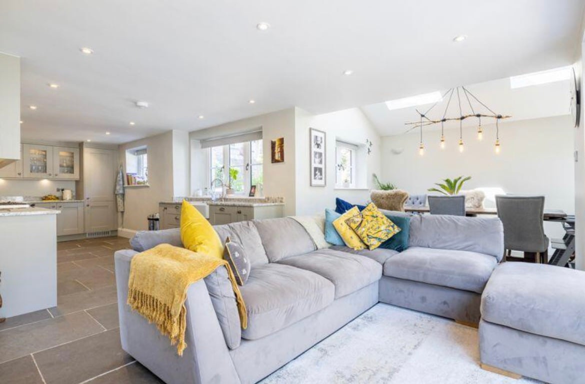 A spacious living area is shown with a large, comfortable sectional sofa adorned with various textured cushions. Natural light filters through multiple windows, highlighting the open design that connects the living space to the kitchen area. A dining table is visible in the background.