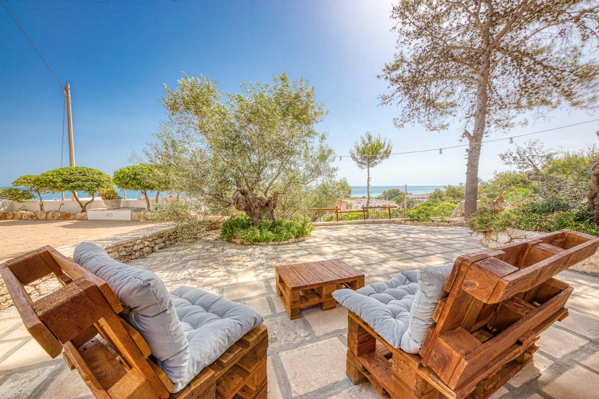 An outdoor seating area is presented, featuring two wooden chairs with blue cushions, positioned on a stone-paved surface. In the background, a view of the sea is visible, framed by trees and shrubs, creating a serene and inviting space.