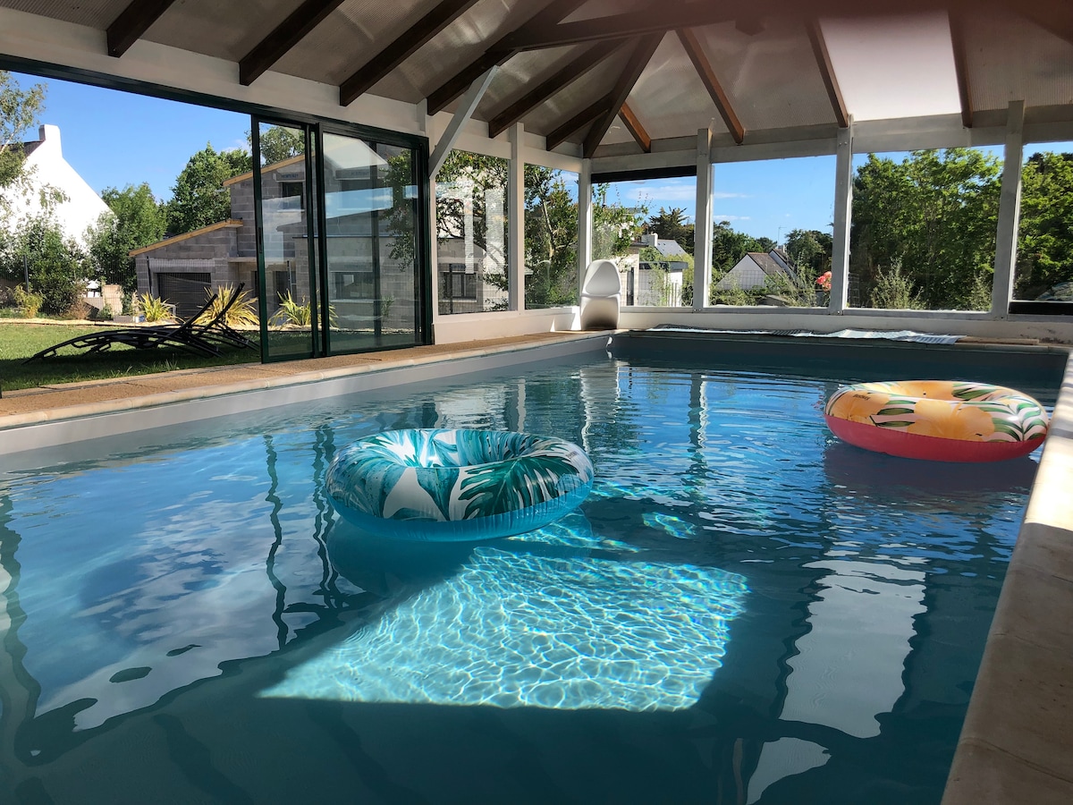 An indoor pool is featured within a spacious area, surrounded by large glass walls that offer views of the outdoor garden. Two colorful pool floats rest on the water's surface, reflecting the sunlight that filters through the ceiling. Natural light enhances the inviting atmosphere.
