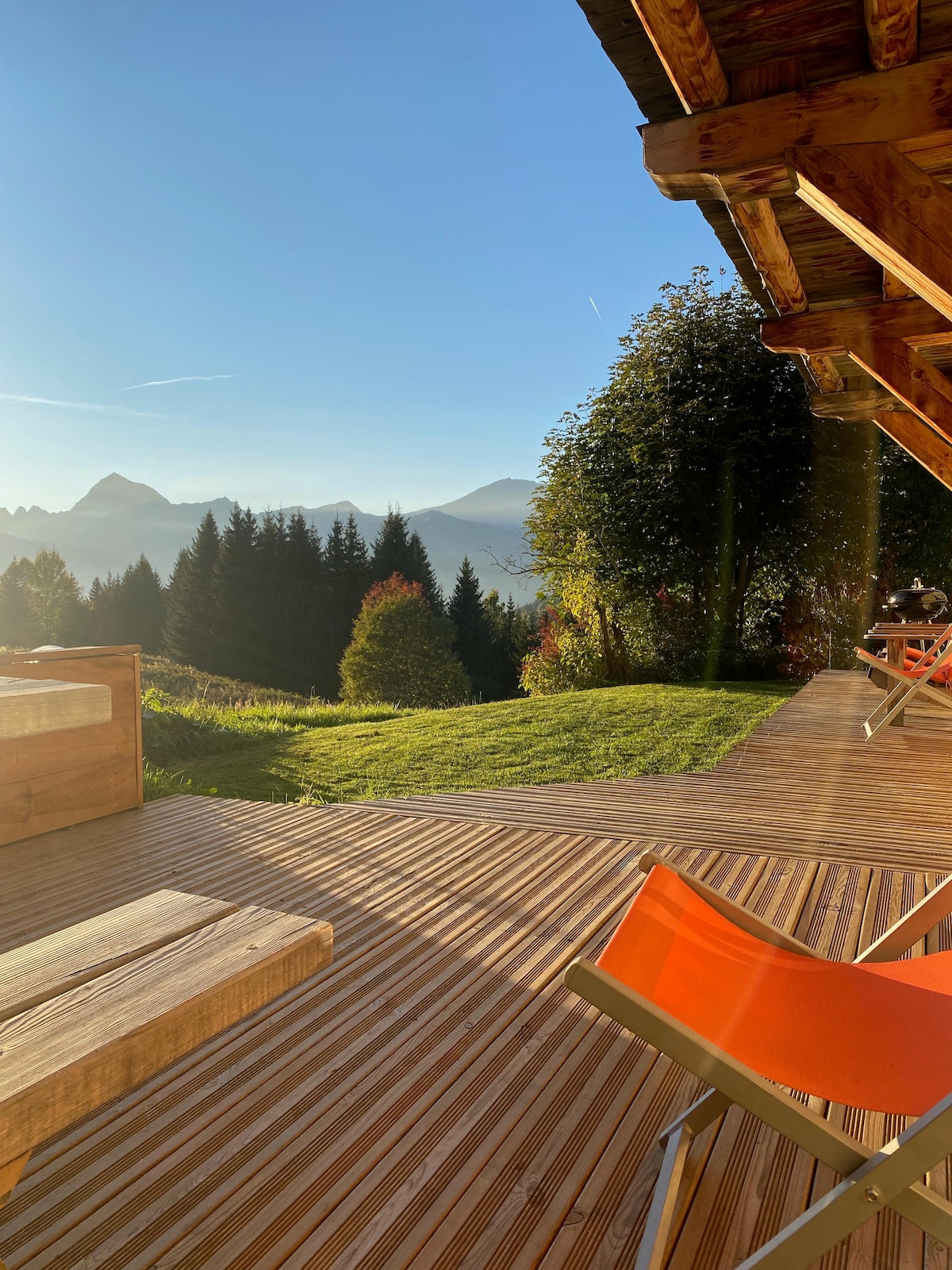 A spacious outdoor deck features wooden flooring and seating options. Lush greenery and trees surround the area, with distant mountain peaks visible under a clear blue sky. Warm sunlight enhances the tranquil atmosphere of the setting.