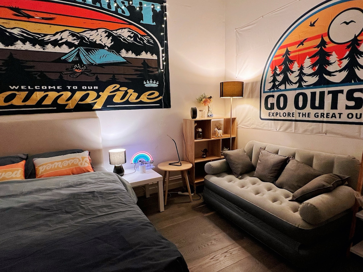 A cozy studio area features a spacious bed with soft bedding; colorful wall tapestries create a campfire theme. A comfortable sofa is positioned nearby, complemented by a small side table with a lamp. Natural light is filtered through the decorated walls.