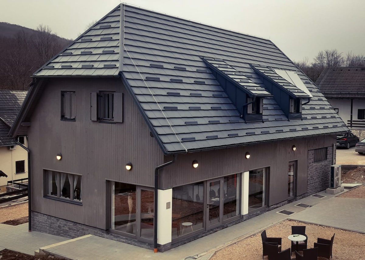 A modern building is showcased, featuring a combination of wooden and stone facades. Large windows and skylights enhance natural light within the structure. Outdoor seating is provided in front, with a paved pathway leading to the entrance.