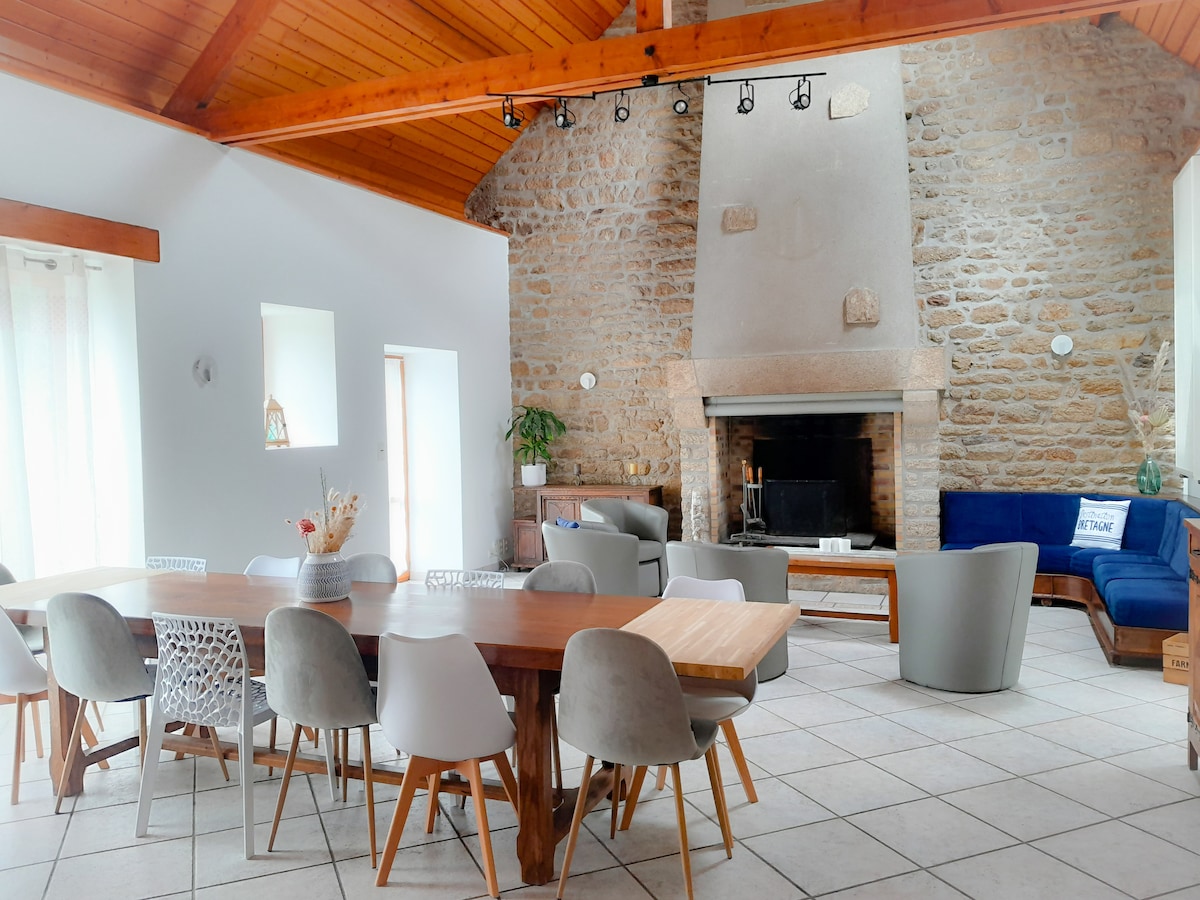 The spacious living area features high vaulted ceilings and a prominent stone fireplace. A large wooden dining table is surrounded by various chairs. A blue sofa provides additional seating, complemented by light streaming through multiple windows.