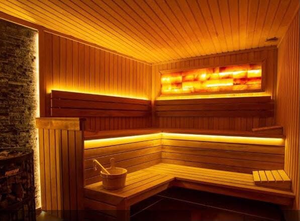 The sauna area is lined with wooden panels, providing a warm and calming environment. Soft, ambient lighting highlights the seating and walls, while a bowl and ladle are positioned nearby for immersion. A stone wall contrasts with the smooth wood surfaces.