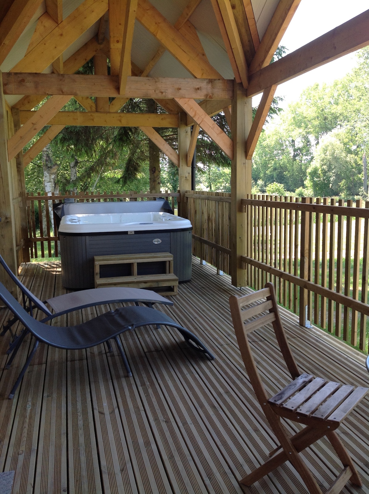 A spacious wooden terrace is fitted with a five-person spa and two lounge chairs, providing a relaxing outdoor space. Lush greenery is visible beyond the railing, offering a serene view of the surrounding bocage.