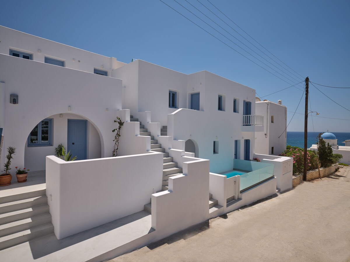 A whitewashed building designed in a Cycladic style features a series of terraces and steps leading to a communal pool. Clear blue sky is visible above, and the sea can be seen in the background, enhancing the serene atmosphere of the location.