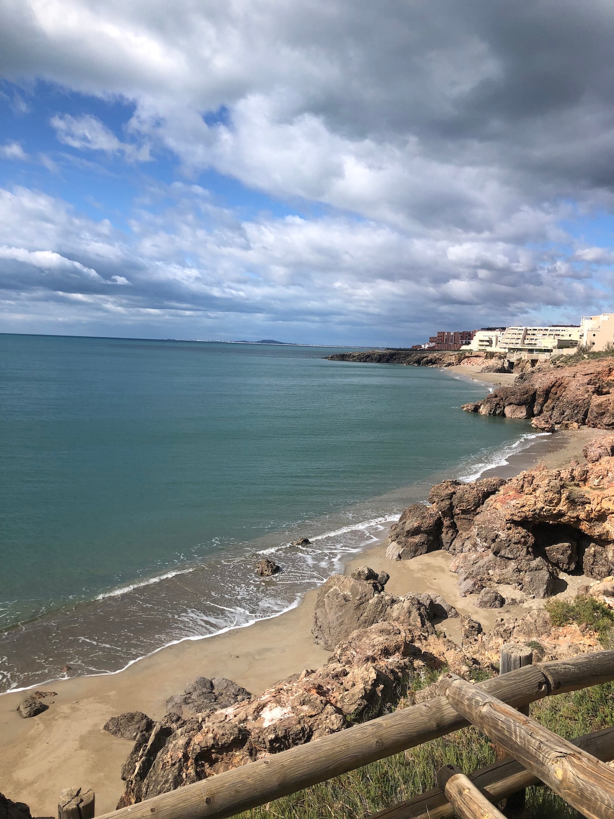 A scenic view of a sandy beach bordered by rocky formations. The calm water glistens under a partly cloudy sky, while a coastal structure can be seen in the background, blending into the coastal landscape.