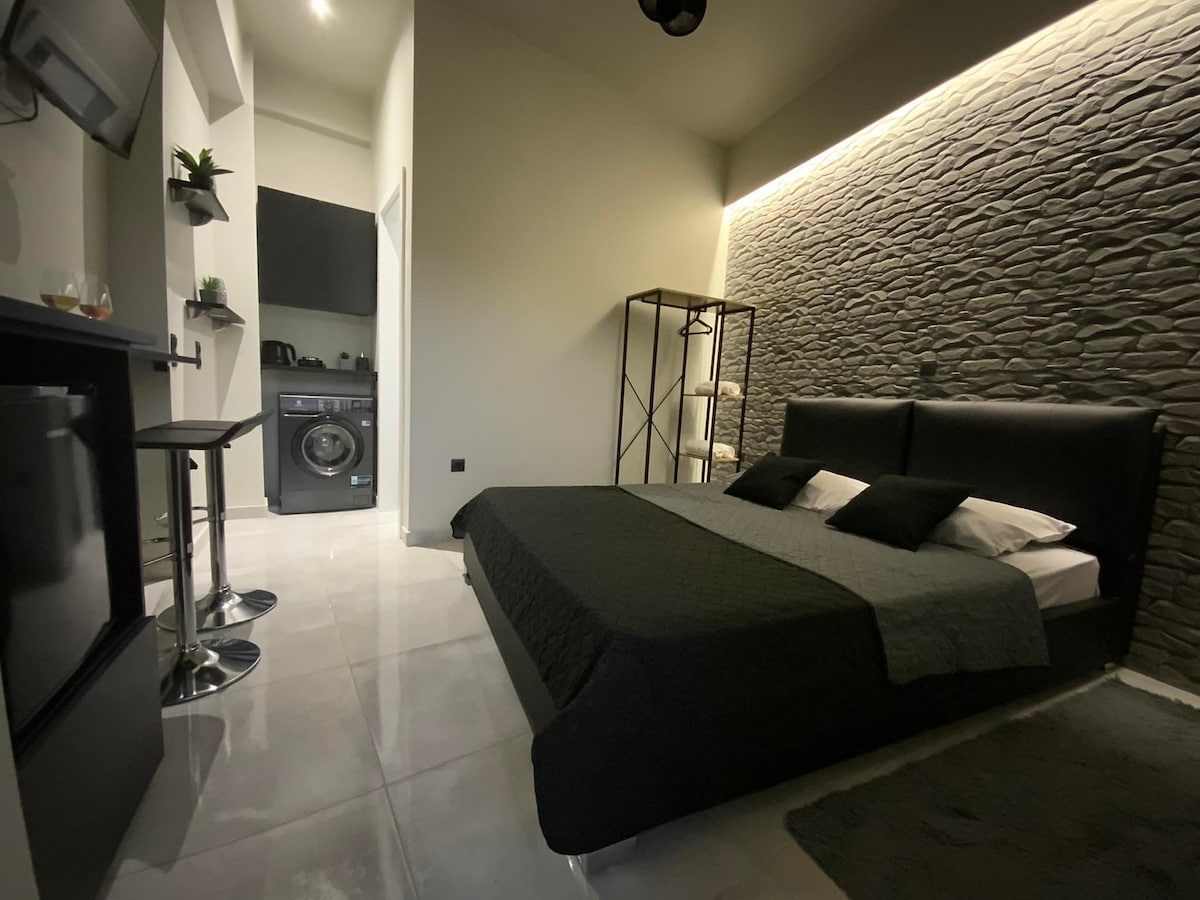A modern mini room features a spacious black bed with dark bedding and contrasting white pillows. A textured stone wall adds depth, while the kitchenette and washer are visible in the background. A small dining area with sleek stools complements the overall design.