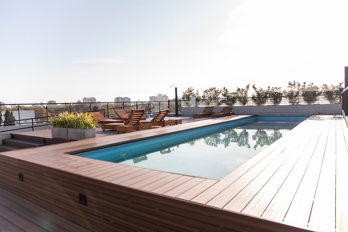 An outdoor pool area is surrounded by wooden decking and landscaped planters. Several lounge chairs are positioned for sunbathing. The pool reflects the clear sky, with city views visible in the background.