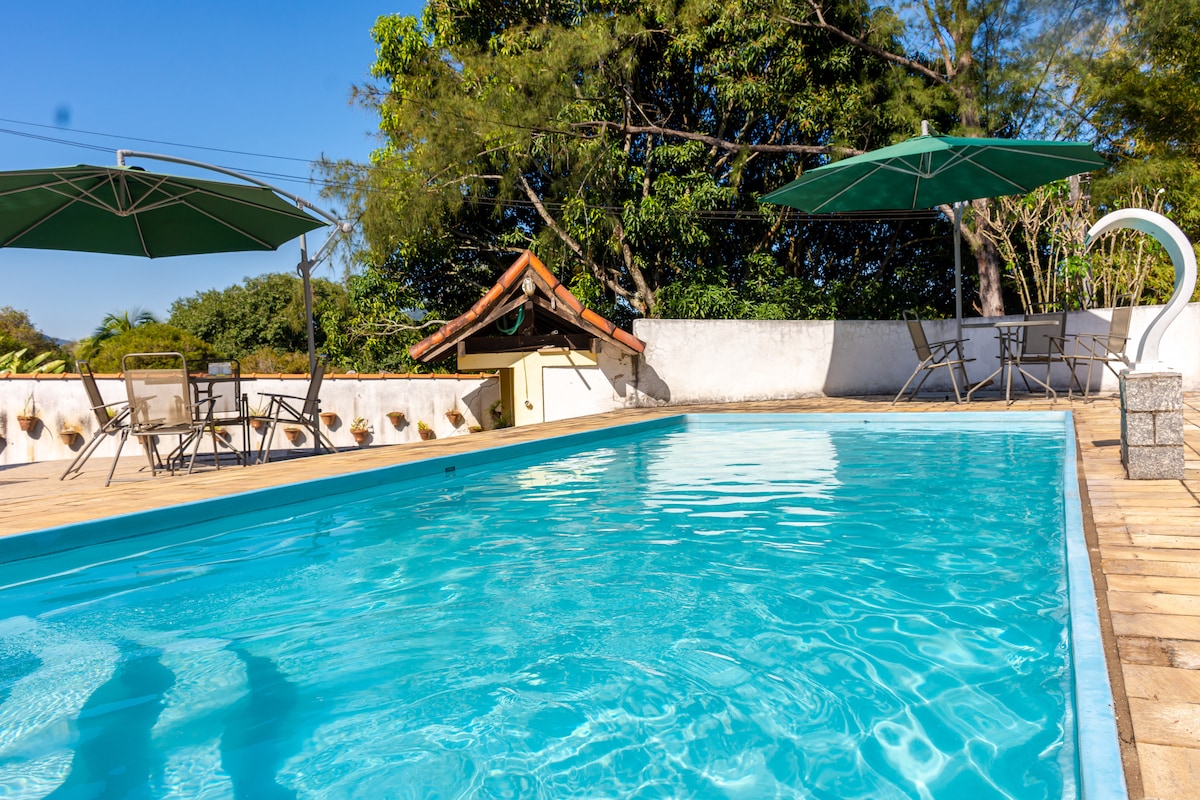 A refreshing swimming pool is surrounded by a sun deck, featuring green umbrellas for shade. Adjacent seating areas include metal chairs and tables, offering a comfortable space for relaxation in a serene natural setting.