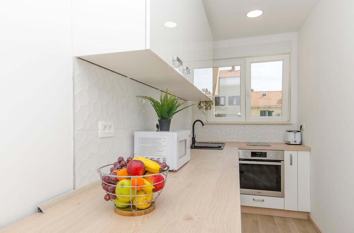 A modern kitchen is presented, featuring light-colored cabinetry and a wooden countertop. A bowl of assorted fresh fruits is showcased prominently. A microwave and oven are integrated within the cabinetry, complemented by a sink and a window allowing natural light to illuminate the space.