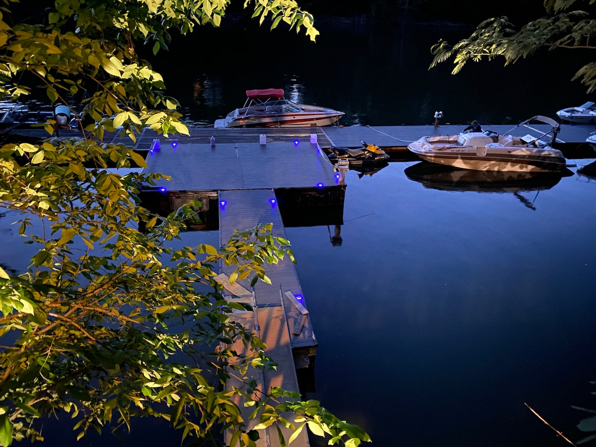 A wooden boat dock extends over a calm body of water, illuminated by blue LED lights. Several boats are moored alongside, surrounded by a serene natural setting with leafy trees framing the scene.