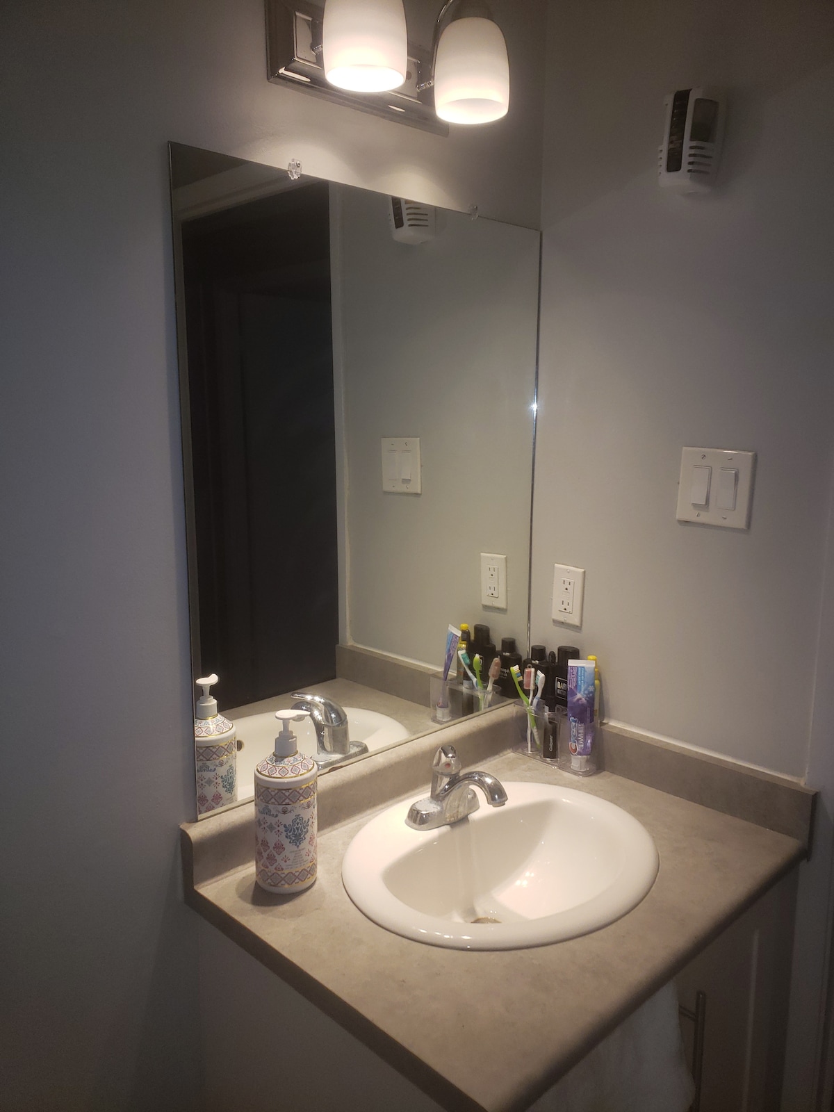 A well-lit bathroom features a large mirror above a smooth countertop with a sink. Personal care items are organized neatly beside a decorative soap dispenser. Soft lighting creates a calm atmosphere in this practical space.