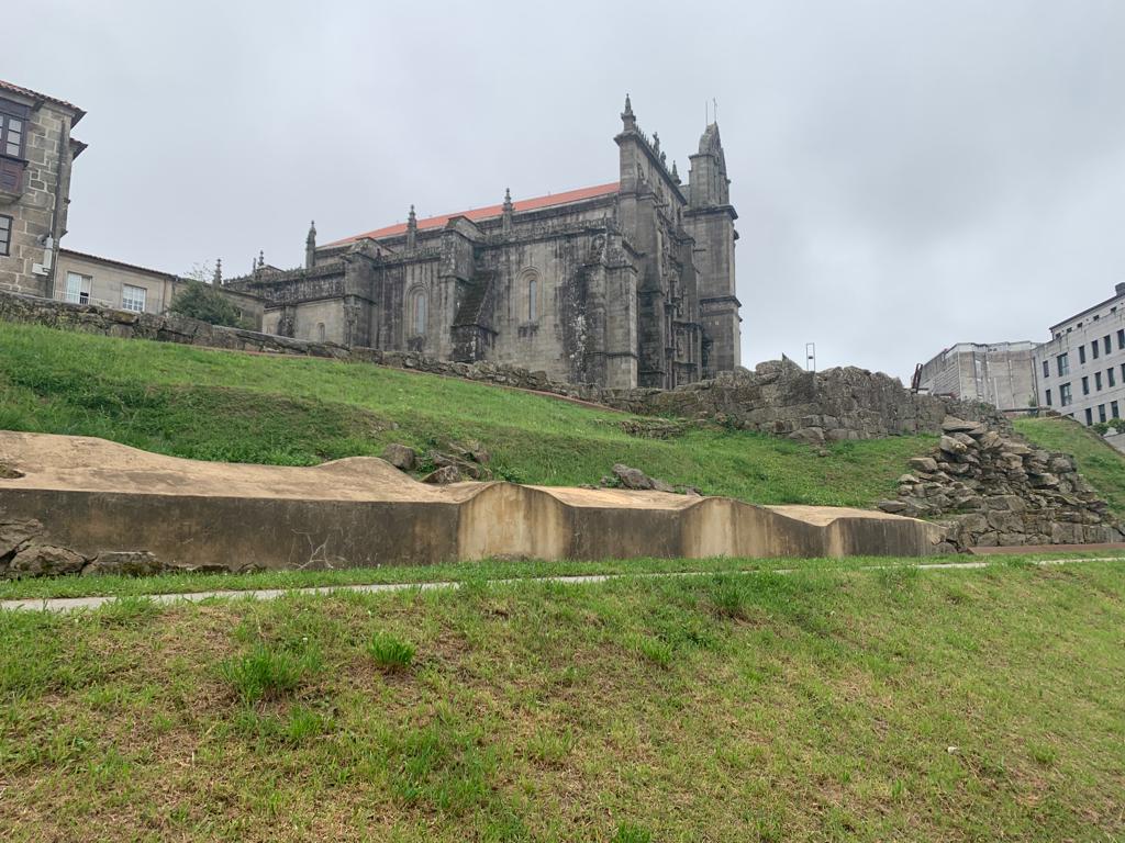 An historic building with intricate stone architecture is situated on elevated ground. Lush green grass surrounds the structure, complemented by rocky outcrops in the foreground. Overcast skies create a muted atmosphere, highlighting the architectural details of the façade.