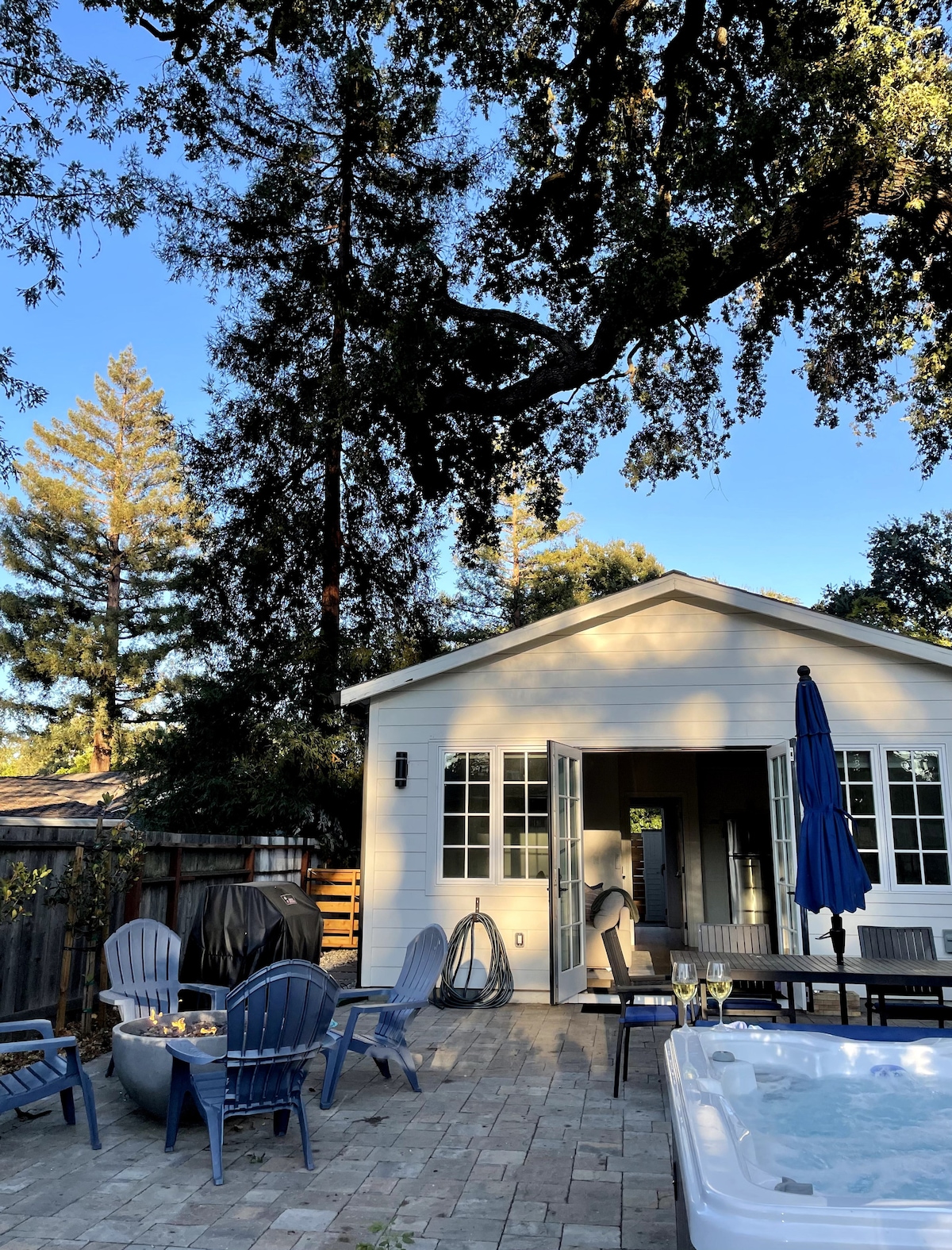 A spacious patio is shown, featuring lounge chairs and a shaded umbrella. The hot tub is positioned nearby, with a view of the charming guest house. Tall trees provide a natural backdrop, and doors are opened to enhance the airy feel of the space.