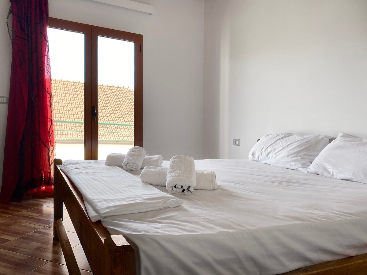 A spacious bedroom features a large bed with crisp white linens and neatly arranged towels. Natural light floods the room through large glass doors, which open to a scenic view. Soft drapes in a rich red add a touch of color to the neutral decor.