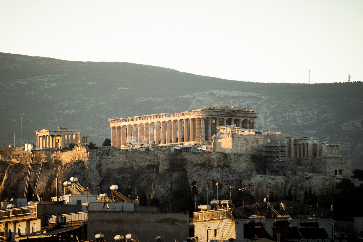 The iconic Acropolis is showcased atop a hill, featuring its ancient columns and structures bathed in warm sunlight. The landscape surrounding it includes rolling hills, while the foreground displays a range of rooftops of nearby buildings.