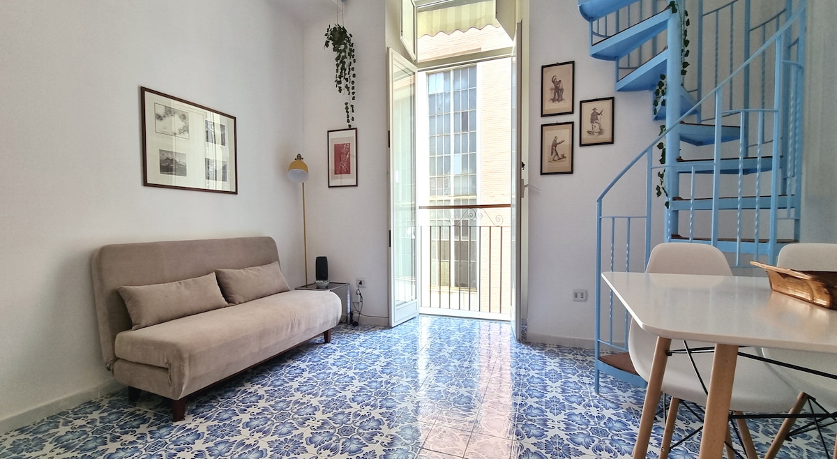 A living area is depicted with a neutral-toned sofa and a white dining table surrounded by chairs. A blue spiral staircase is visible, leading to an upper level. Large windows allow natural light to fill the space, with blue-and-white patterned tiles enhancing the floor.