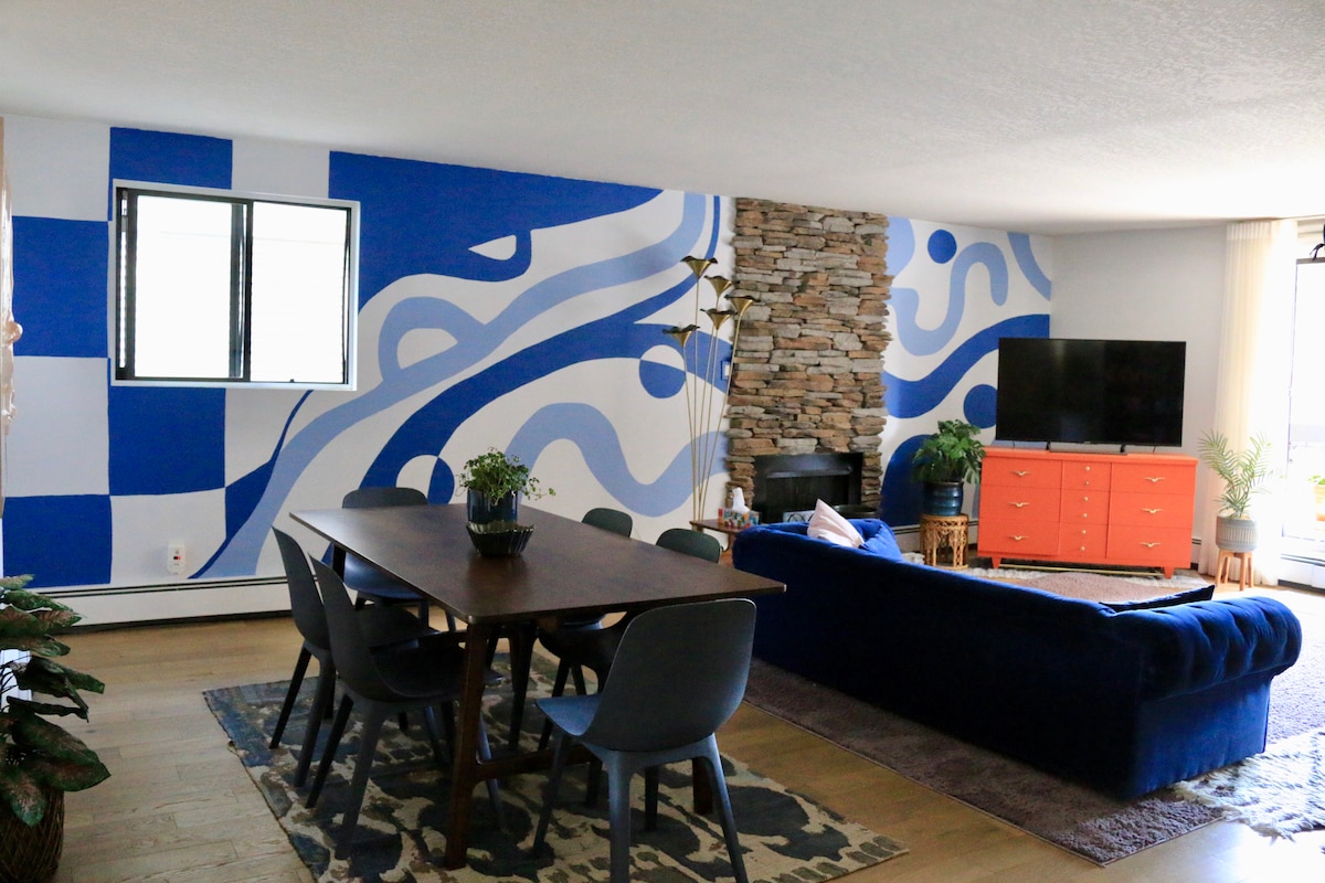 A spacious living area showcases a large blue sectional sofa and a wooden dining table with grey chairs. A stone accent wall complements a colorful mural, while a media console holds a television. Natural light brightens the space through a window.