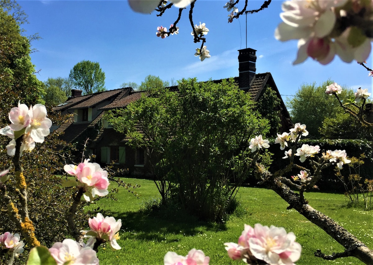 A charming rural home is partially obscured by blooming flowers, surrounded by a lush green garden. The structure features a traditional roof and a prominent chimney. A variety of trees and vegetation create a tranquil setting, enhancing the natural beauty of the outdoor space.